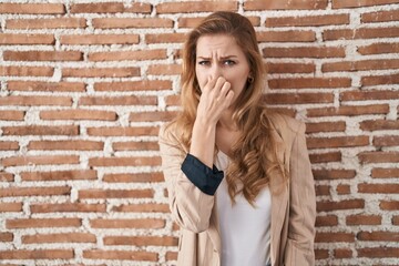 Beautiful blonde woman standing over bricks wall smelling something stinky and disgusting, intolerable smell, holding breath with fingers on nose. bad smell