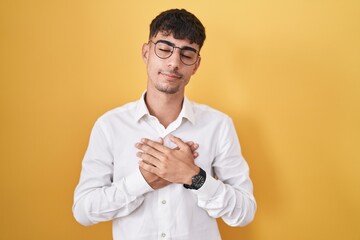 Young hispanic man standing over yellow background smiling with hands on chest with closed eyes and grateful gesture on face. health concept.