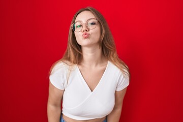 Young caucasian woman standing over red background looking at the camera blowing a kiss on air being lovely and sexy. love expression.