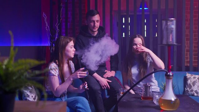 two young women and a young man in a hookah lounge on sofas, laughing, chatting and smoking a hookah. exhale a lot of white smoke