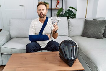 Middle age man with beard wearing arm on sling for motorbike accident holding insurance money...