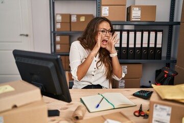 Young hispanic woman working at small business ecommerce shouting angry out loud with hands over mouth