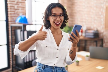 Young hispanic woman working at the office using smartphone smiling happy and positive, thumb up doing excellent and approval sign