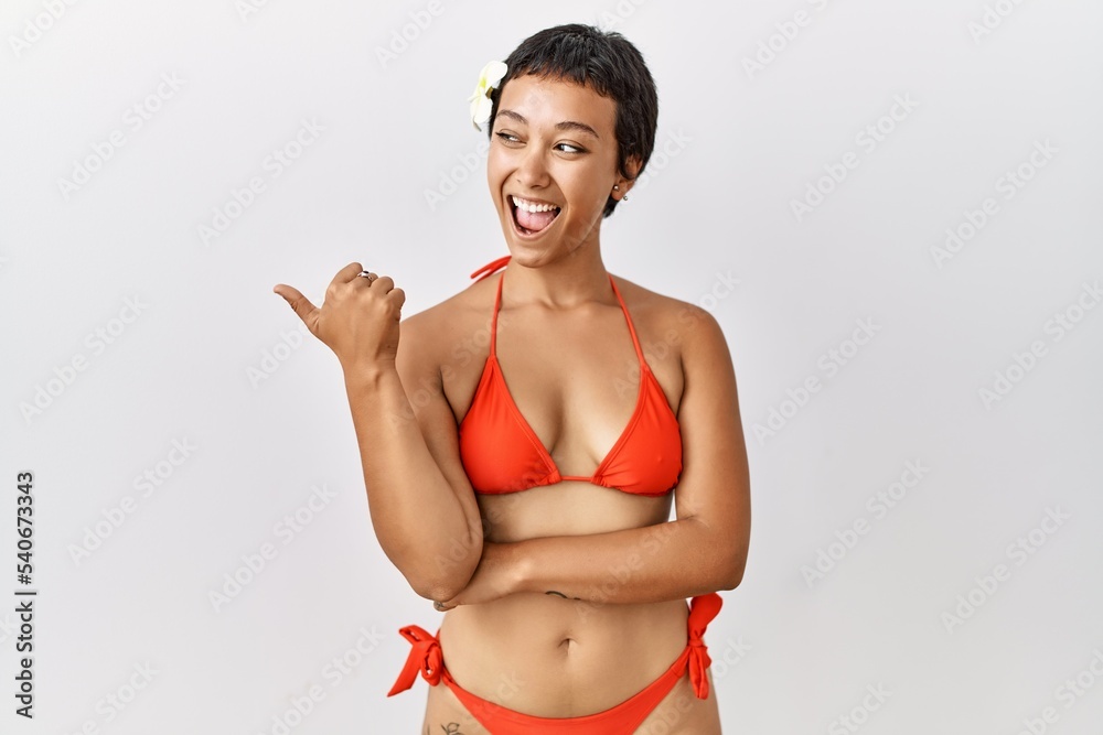 Wall mural Young hispanic woman with short hair wearing bikini smiling with happy face looking and pointing to the side with thumb up.