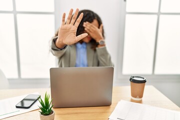 Beautiful hispanic business woman sitting on desk at office working with laptop covering eyes with hands and doing stop gesture with sad and fear expression. embarrassed and negative concept.