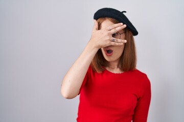 Young redhead woman standing wearing glasses and beret peeking in shock covering face and eyes with...