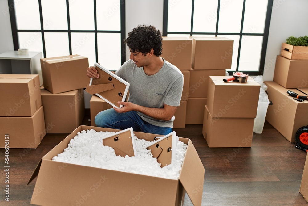 Sticker young hispanic man smiling happy unboxing cardboard box at new home.