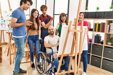 Group of young paint students smiling happy and looking draw of disabled partner sitting on...