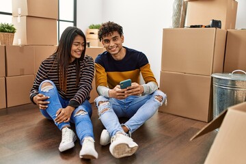 Young latin couple using smartphone sitting on the floor at new home.