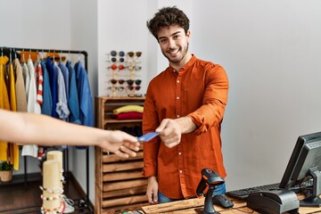 Young hispanic shopkeeper man giving credit card to customer at clothing store.