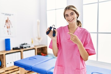 Young blonde woman working at pain recovery clinic holding hand strengthener smiling happy pointing with hand and finger