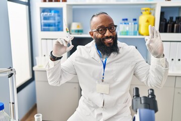 African american man working at scientist laboratory holding syringe doing ok sign with fingers, smiling friendly gesturing excellent symbol
