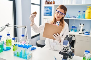 Young blonde woman wearing scientist uniform holding kiwi and clipboard at laboratory