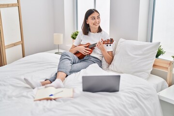 Young hispanic woman playing ukelele sitting on bed at bedroom