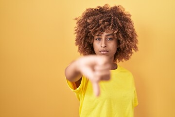 Young hispanic woman with curly hair standing over yellow background looking unhappy and angry...