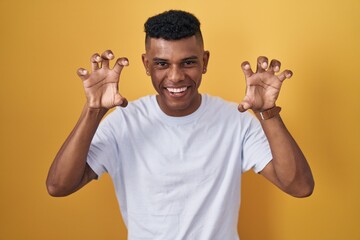 Young hispanic man standing over yellow background smiling funny doing claw gesture as cat,...