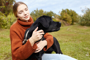 a happy woman hugs her black labrador dog while walking in an autumn park