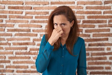 Brunette woman standing over bricks wall smelling something stinky and disgusting, intolerable smell, holding breath with fingers on nose. bad smell
