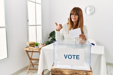 Hispanic business woman voting putting envelop in ballot box pointing to the back behind with hand...