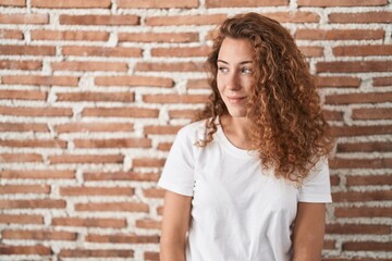 Young caucasian woman standing over bricks wall background looking away to side with smile on face, natural expression. laughing confident.