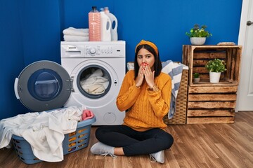 Young hispanic woman doing laundry laughing and embarrassed giggle covering mouth with hands, gossip and scandal concept