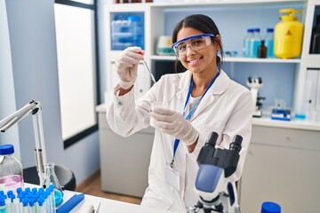 Young latin woman wearing scientist uniform using pipette at laboratory
