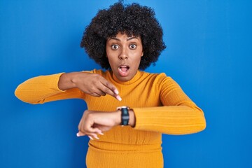 Black woman with curly hair standing over blue background in hurry pointing to watch time,...
