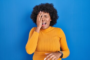 Fototapeta na wymiar Black woman with curly hair standing over blue background yawning tired covering half face, eye and mouth with hand. face hurts in pain.