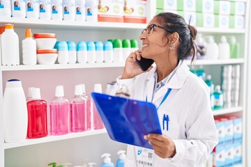 Young beautiful hispanic woman pharmacist talking on smartphone reading document at pharmacy