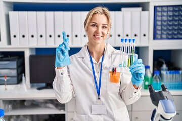 Young caucasian woman working at scientist laboratory holding test tubes smiling with an idea or question pointing finger with happy face, number one