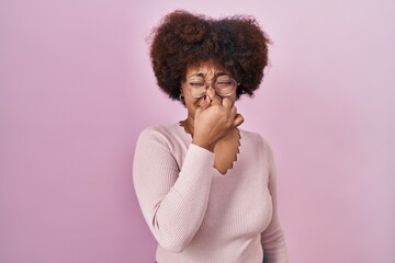 Young african american woman standing over pink background smelling something stinky and disgusting, intolerable smell, holding breath with fingers on nose. bad smell