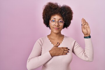 Young african american woman standing over pink background swearing with hand on chest and open...