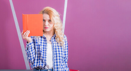 Young girl blonde stands background of pink wall, holds an open book in front of her and looks with a strict look at camera.