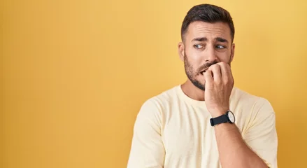 Fotobehang Handsome hispanic man standing over yellow background looking stressed and nervous with hands on mouth biting nails. anxiety problem. © Krakenimages.com