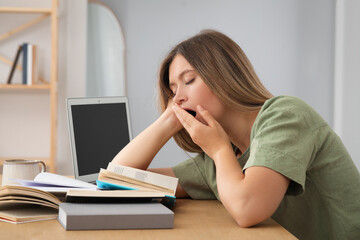 Sleepy young woman studying at wooden table indoors