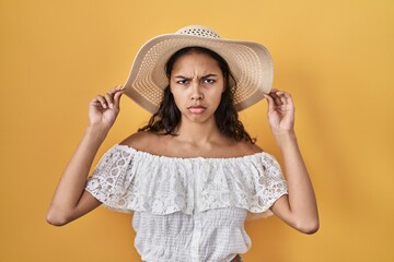 Young brazilian woman wearing summer hat over yellow background skeptic and nervous, frowning upset because of problem. negative person.