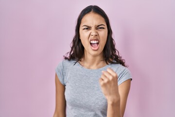 Young brazilian woman wearing casual t shirt over pink background angry and mad raising fist...