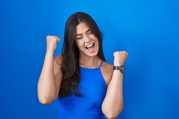 Fototapeta na wymiar Hispanic woman standing over blue background celebrating surprised and amazed for success with arms raised and eyes closed. winner concept.