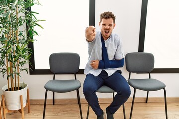 Handsome young man sitting at doctor waiting room with arm injury pointing displeased and frustrated to the camera, angry and furious with you