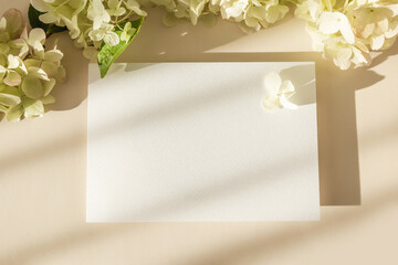 Invitation card mockup with hydrangea flowers on beige pastel background. Template blank of white paper mock up for branding and advertising. Top view, flat lay, copy space.