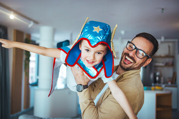 Dream big. Loving father helps his son fly like a superhero. Boy play fly with his dad at home. Cheerful familyare having fun together. Father holding son in superhero costume flying at home