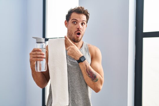 Young hispanic man wearing sportswear drinking water surprised pointing with finger to the side, open mouth amazed expression.