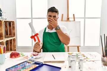 Young hispanic man at art studio holding degree laughing and embarrassed giggle covering mouth with...