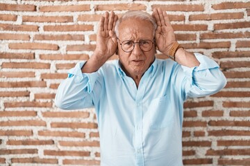 Senior man with grey hair standing over bricks wall doing bunny ears gesture with hands palms...