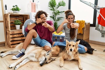 Young hispanic couple doing laundry with dogs smiling doing talking on the telephone gesture and...