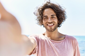 Young hispanic man smiling happy make selfie by the camera standing at the beach.