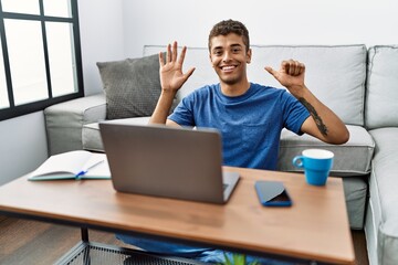 Young handsome hispanic man using laptop sitting on the floor showing and pointing up with fingers number six while smiling confident and happy.