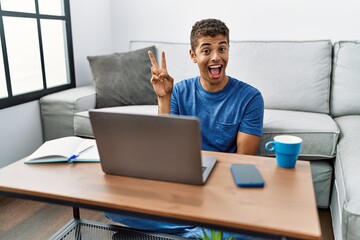 Young handsome hispanic man using laptop sitting on the floor smiling with happy face winking at the camera doing victory sign. number two.