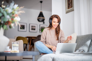 Beautiful woman at home sitting on the sofa while using laptop and headphone - Powered by Adobe