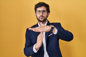 Handsome latin man standing over yellow background doing time out gesture with hands, frustrated...